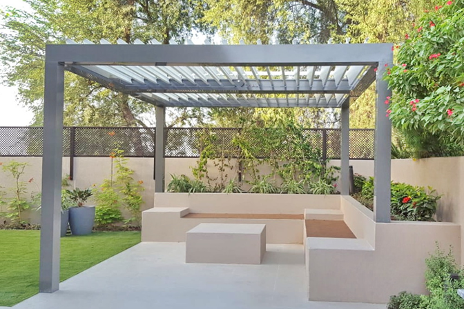 A gray louvered pergola over a detached patio with an L-shaped concrete bench and a matching concrete table in front of it and trees in the background. 